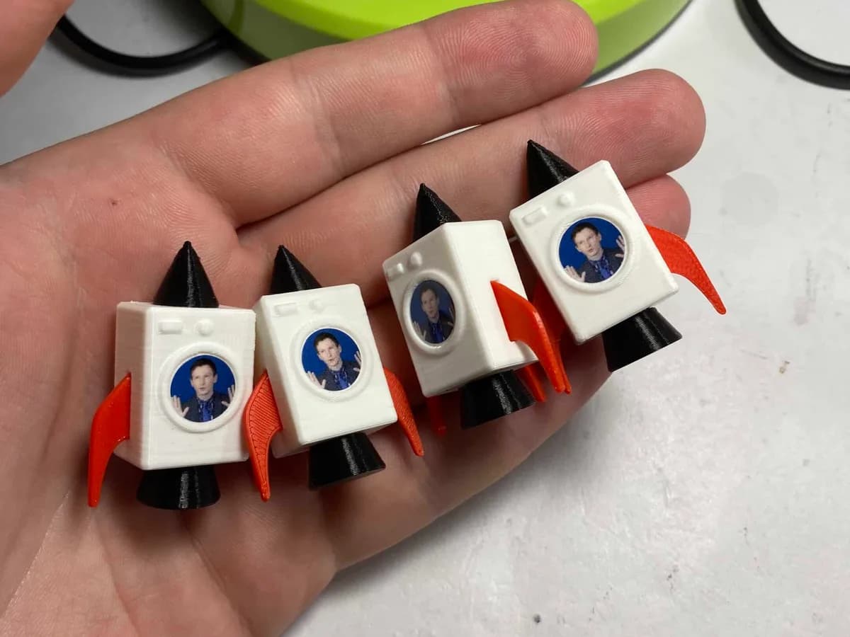 Čtyři hotové figurky raketopraček v dlani — bílé tělo, černý kužel a oranžová křídla.